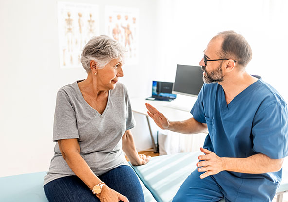 An orthopedic surgeon is sitting next to a  senior woman in an exam room. He is explaining something to her.